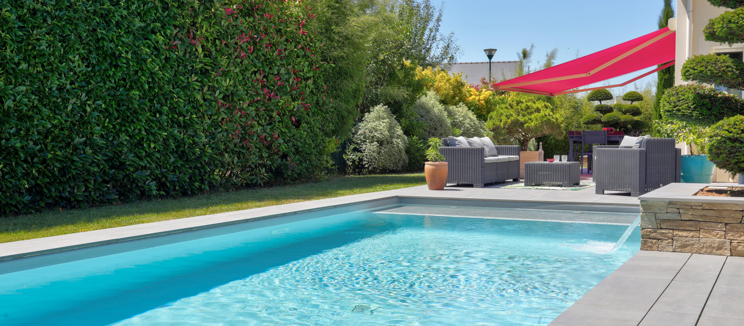 Piscine rectangulaire turquoise avec terrasse en dalles grises, salon de jardin et haie