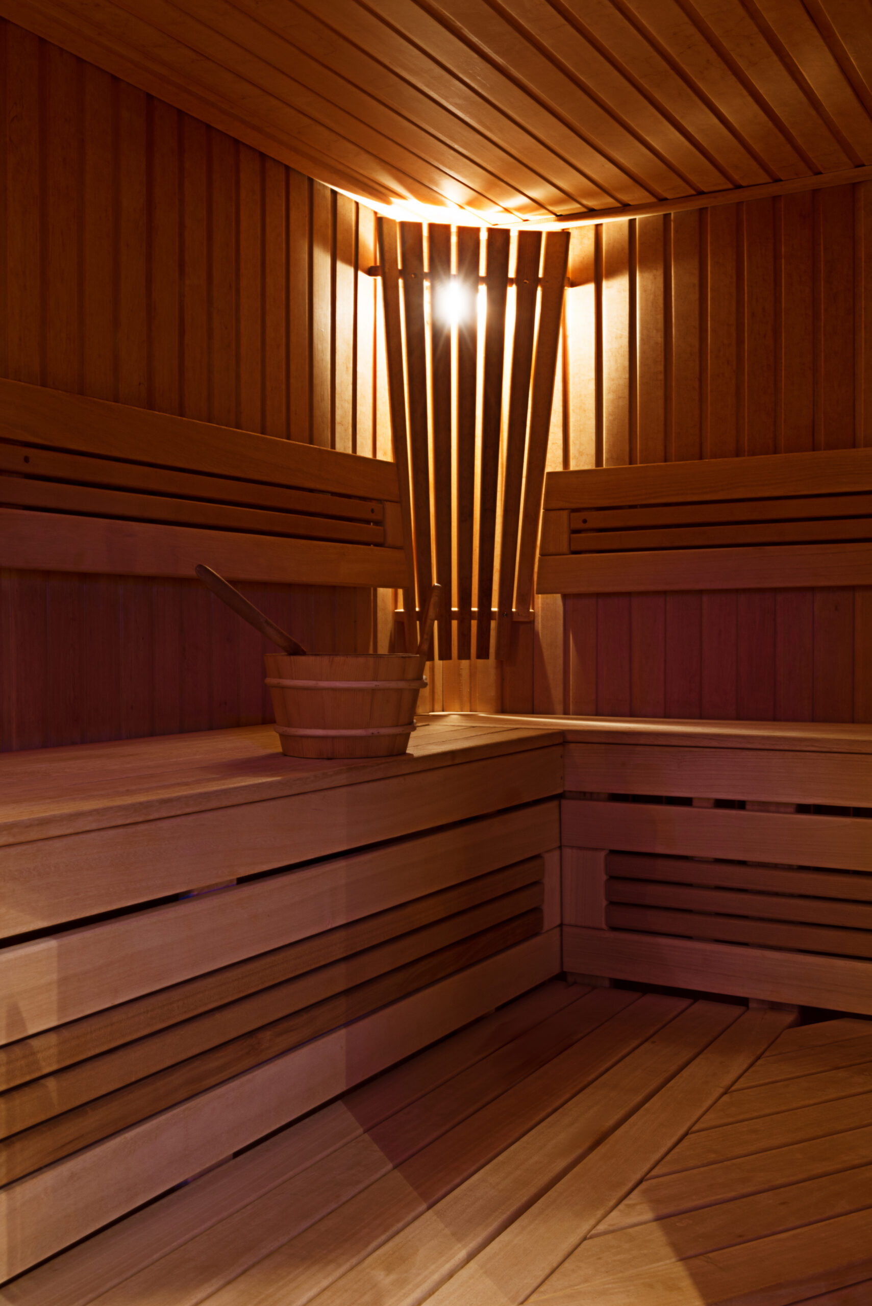 Intérieur d’un sauna en bois clair avec bancs en L, éclairage d’ambiance et seau traditionnel